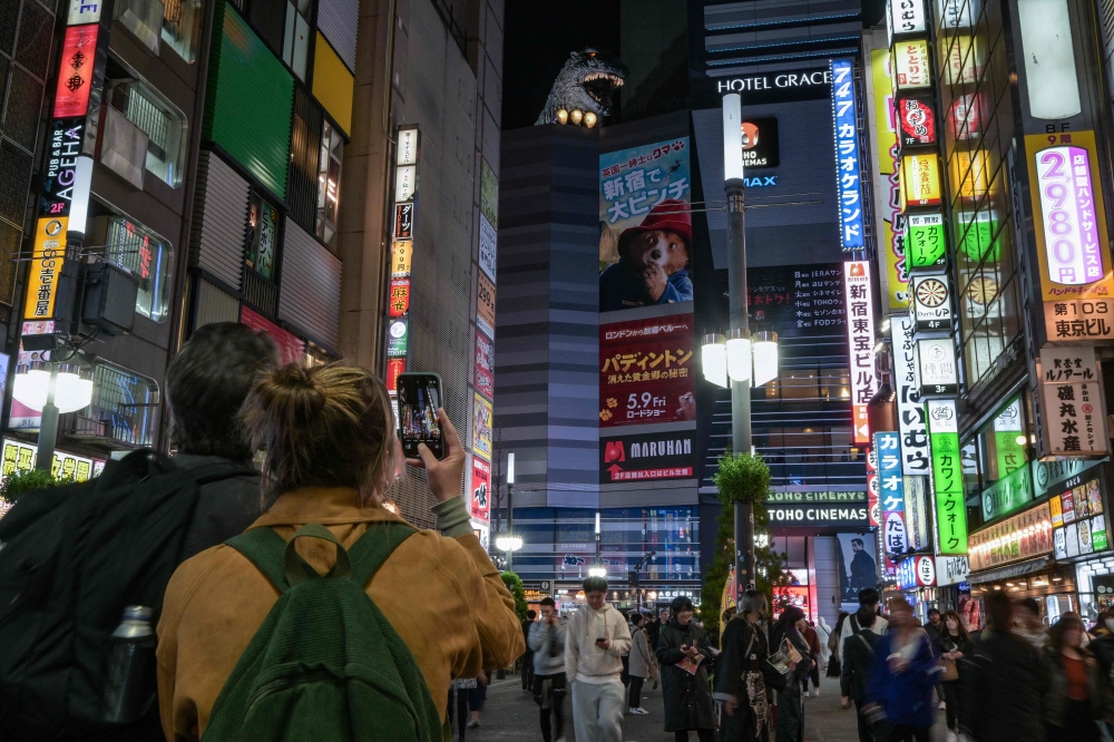 272333 This photo taken on April 8, 2025 shows a tourist (left) taking a photo of a figure of classic film monster Godzilla (top centre) pictured at the top of a building in the red-light entertainment area of Kabukicho in the Shinjuku district of Tokyo. — AFP pic
